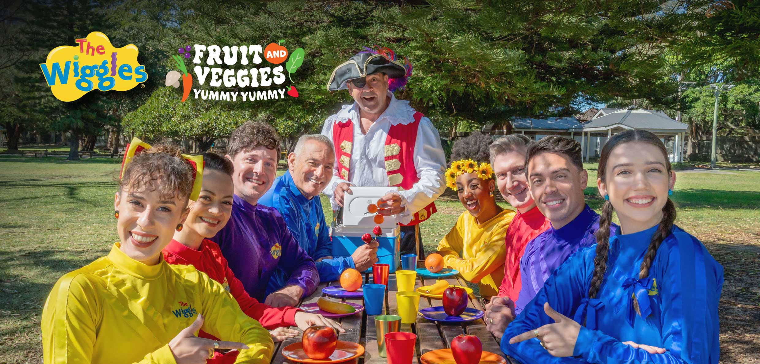 A group of nine people in colorful outfits sitting at a picnic table with fruits and a person in a pirate costume in the background.