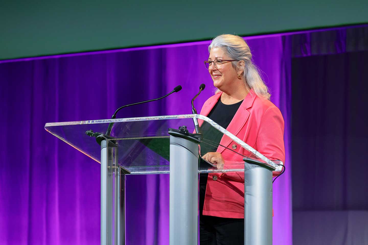 Brenda Briggs in a pink blazer speaks at a clear podium with a purple-lit background.