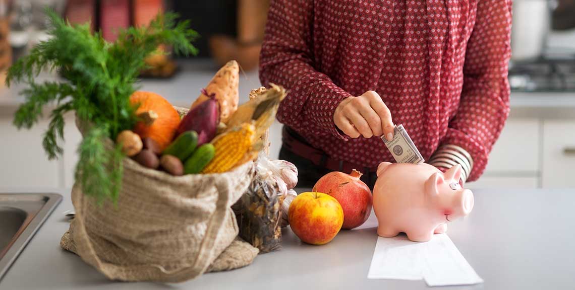 Person placing a dollar bill into a pink piggy bank beside a sack of fresh vegetables and fruit on a kitchen counter.
