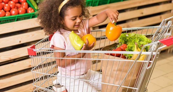 Young child in a shopping cart holding bell peppers in a grocery store.