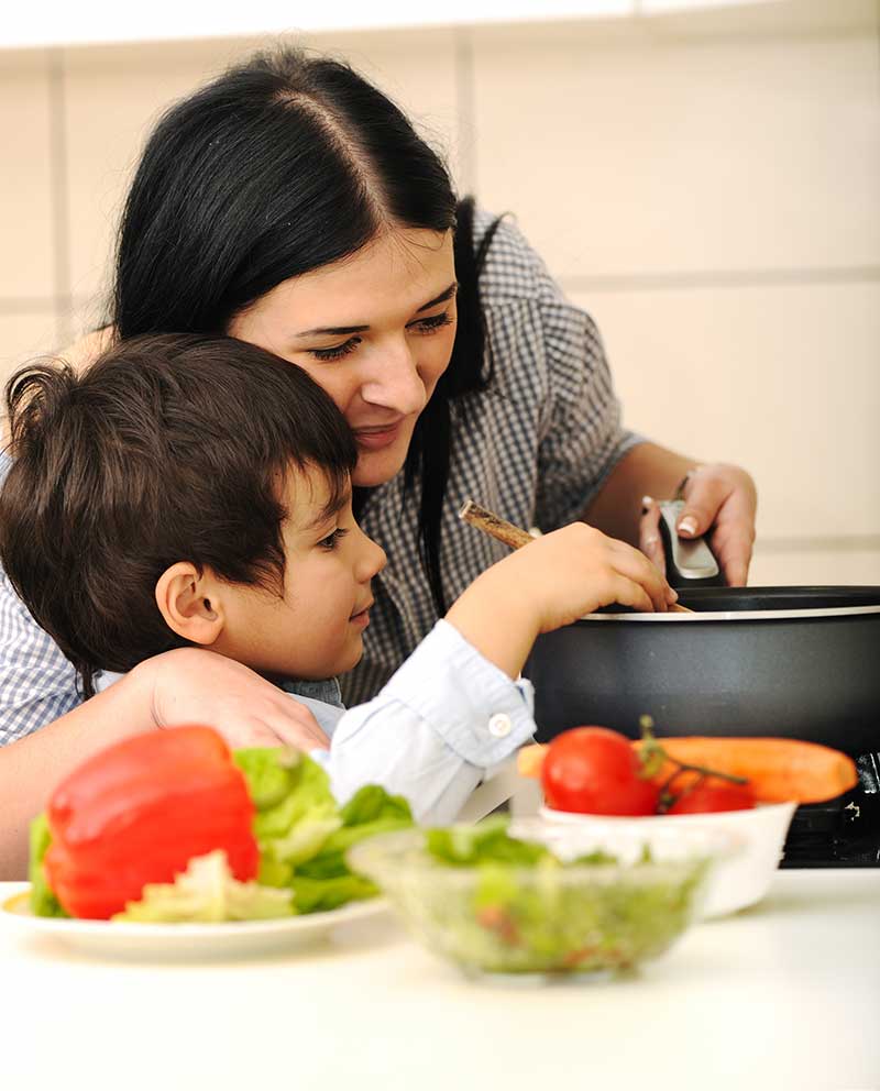 Woman and child cooking together in a kitchen, surrounded by fresh vegetables.