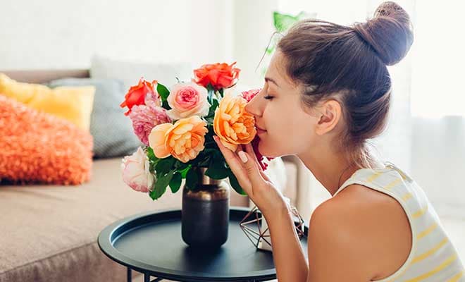 CMS ALT TEXT A woman in her living room gently holds a peach colored flower in her hand and smells the flower with her eyes closed.
