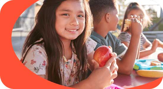 Three children sitting at an outdoor table, with a girl in the foreground holding an apple and smiling.