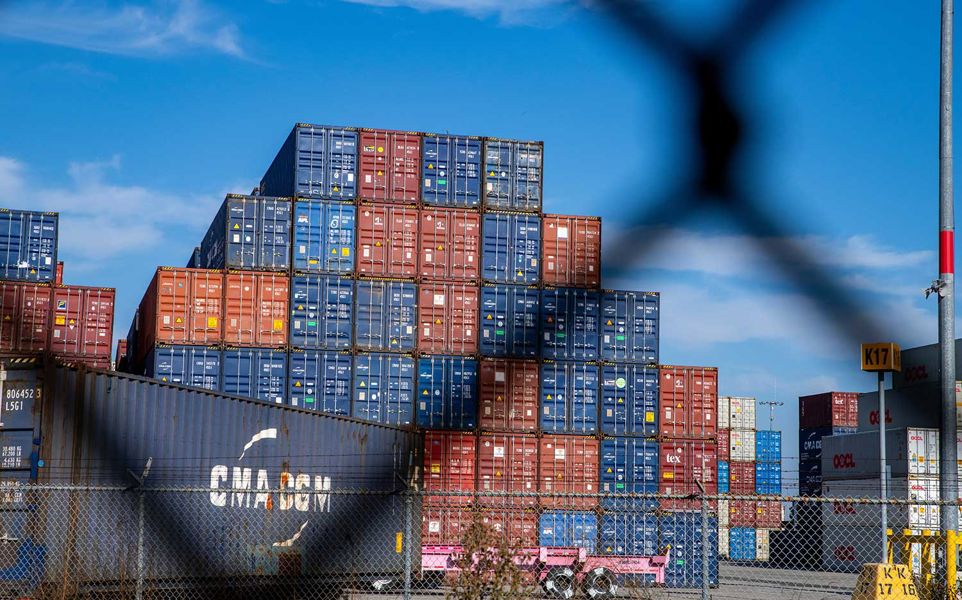 A stack of red, blue, and white shipping containers in a yard, seen behind a blurry chain-link fence, under a blue sky.