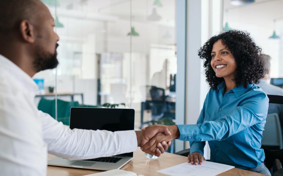 A woman shakes hands with a man who is about to interview her. She sits with her resume infrong of her looking at the man while shaking his hand. 