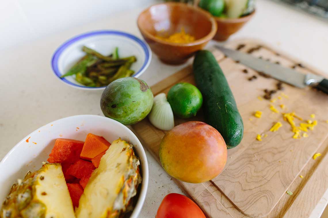 A kitchen counter with sliced pineapple, papaya, mango, lime, onion, cucumber, tomato, and bowls of cheese and green beans.