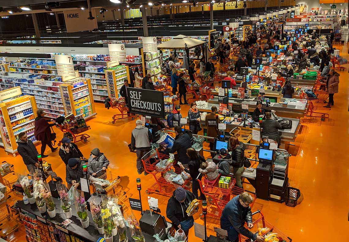 A busy grocery store with crowded check-out counters and self-service stations, featuring an orange floor and shelves stocked with goods.