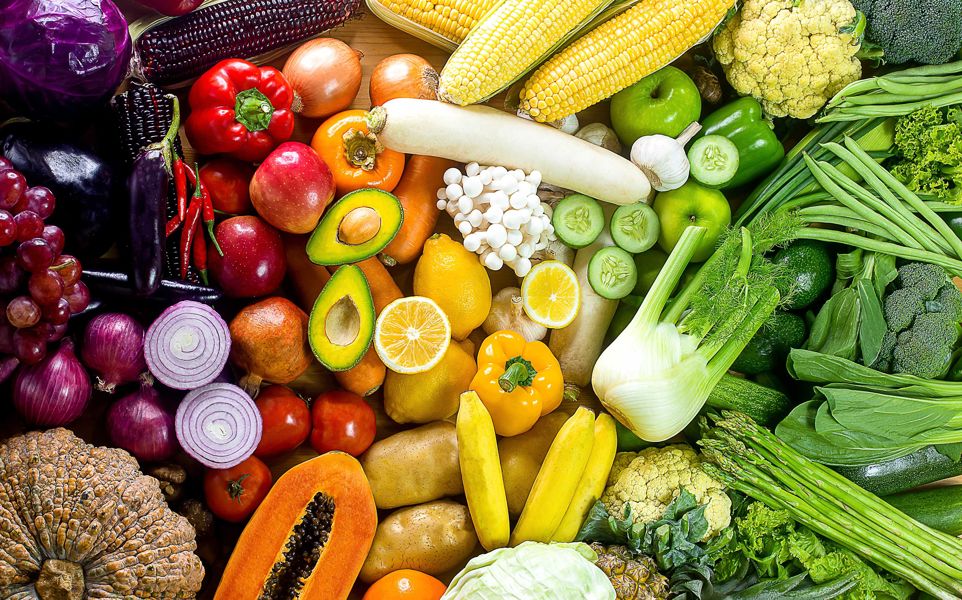 Fresh colorful vegetables and fruits spread out on a table including: purple cabbage, red peppers, avocado, papaya, fennel, tomato, red onion and more. 
